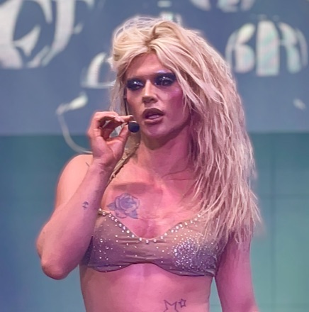 Bimini performing on stage in Torquay in 2021, long blonde hair, in a sequinned bra top, holding a headset microphone.