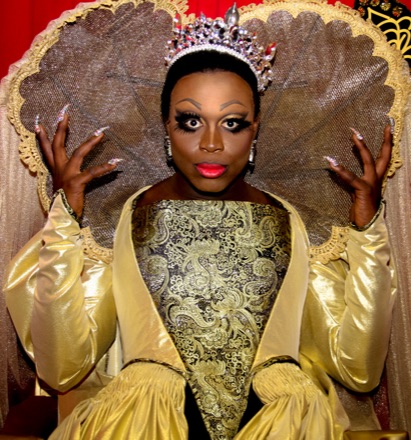 Bob the Drag Queen in drag at RuPaul's DragCon in 2017, wearing a jewelled crown and a gold brocade gown, posed before a lace-edged throne.