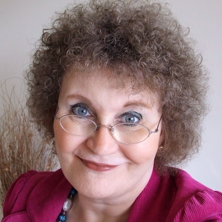 Christine Burns in close-up in 2010, smiling, with curly hair and round glasses, in a magenta top.