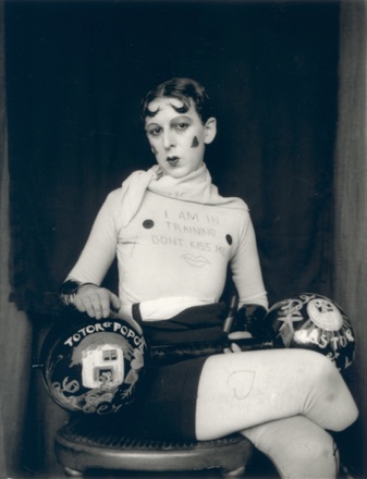 Claude Cahun's self-portrait “I am in training, don't kiss me”: shaved head, painted-on curls and heart-shaped cheeks, in a costume marked with the title, holding a pair of decorated dumbbells.