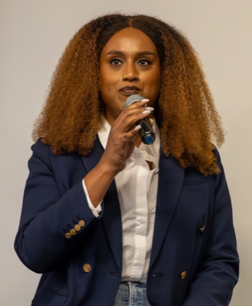 Honey Mahogany speaking into a microphone, with long curly hair, in a navy blazer over a white shirt.
