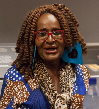 Janetta Johnson speaking on a panel in San Francisco in 2018, with long twisted hair and red glasses, in a patterned blue jacket and a leopard-print scarf.