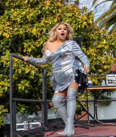 Jiggly Caliente performing on an outdoor stage at the 2023 San Francisco Trans March, in a silver sequinned dress and thigh-high silver boots.