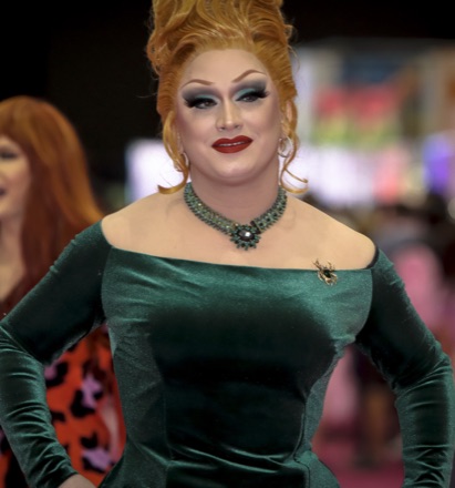 Jinkx Monsoon in drag at RuPaul's DragCon in 2022, red hair piled high, in an off-the-shoulder green velvet gown and a jewelled choker.
