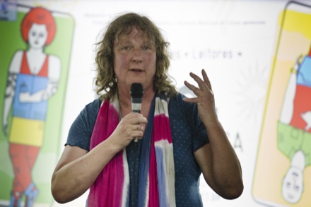 Jo Clifford speaking into a microphone at a literary festival in Rio de Janeiro in 2016, with curly hair and a bright pink striped scarf.