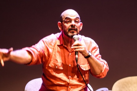 João W. Nery speaking on stage in São Paulo, bald and bearded, in glasses and an orange shirt, holding a microphone.