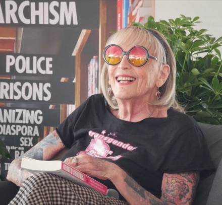 Kate Bornstein laughing, in round tinted glasses and a black T-shirt, with a book in her lap and shelves of books behind her.