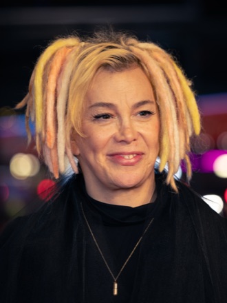 Lana Wachowski at the Berlinale in 2025, smiling, with shoulder-length blonde and orange dreadlocks, in a black high-necked top and a pendant.