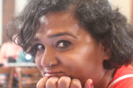 Living Smile Vidya in close-up, chin resting on her hands, with short curly hair and a nose stud.