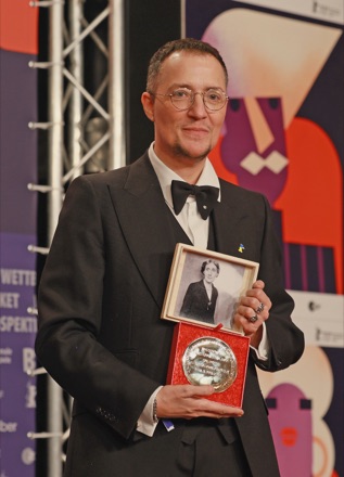 Paul B. Preciado in a dinner jacket and bow tie at the Berlinale, holding the jury award and a framed portrait photograph.