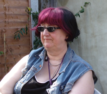 Roz Kaveney sitting outdoors in 2007, with a magenta-streaked bob and dark glasses, in a denim waistcoat.