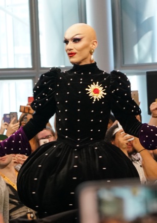 Sasha Velour in drag at DragCon in 2017, bald, in a black high-necked gown studded with pearls and a jewelled brooch, in front of a crowd.