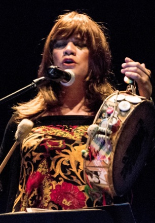 Susy Shock singing on stage in 2013, with long brown hair, in a floral top, holding a hand drum.